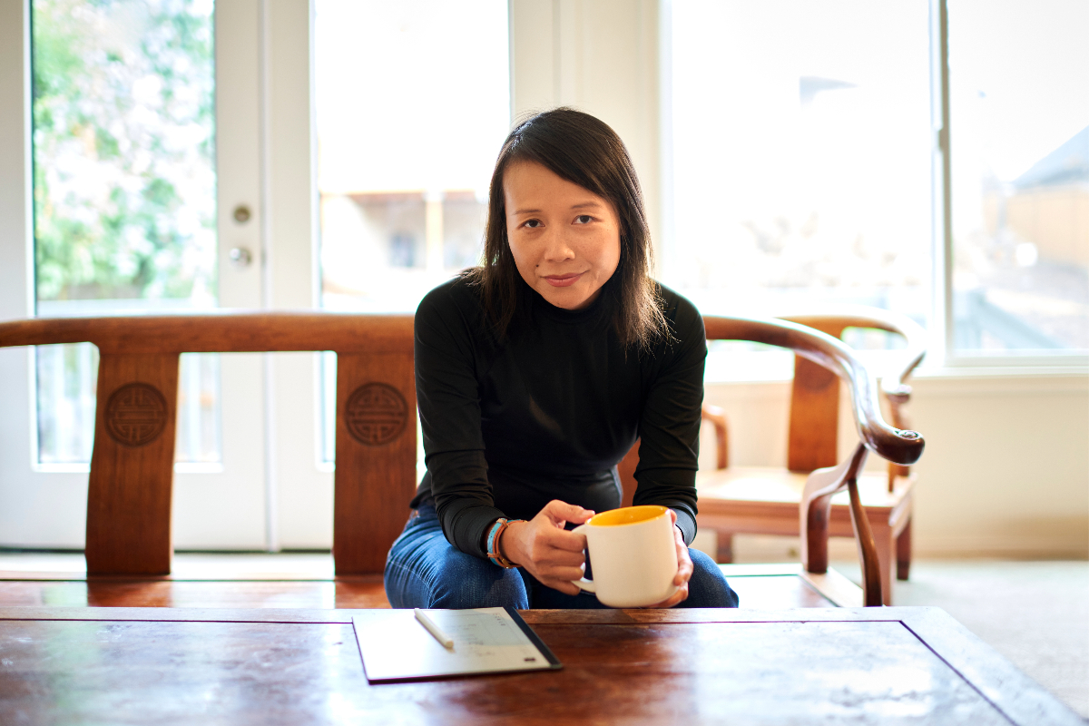 Julie Pham sits in her living room, in a black, turteneck and jeans, hugging a mug of coffee, and leaning foward. In front of her on a large coffee table is a pad of paper and pen.