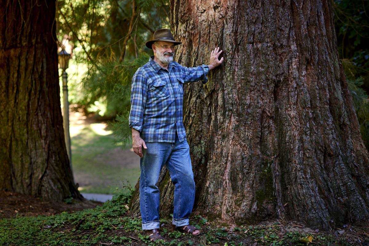 Michael Mason is standing next to tree that is several times his circumference. He's wearing blue jeans, a blue flannel shirt, and a hat, and one hand is resting against the tree to his left.