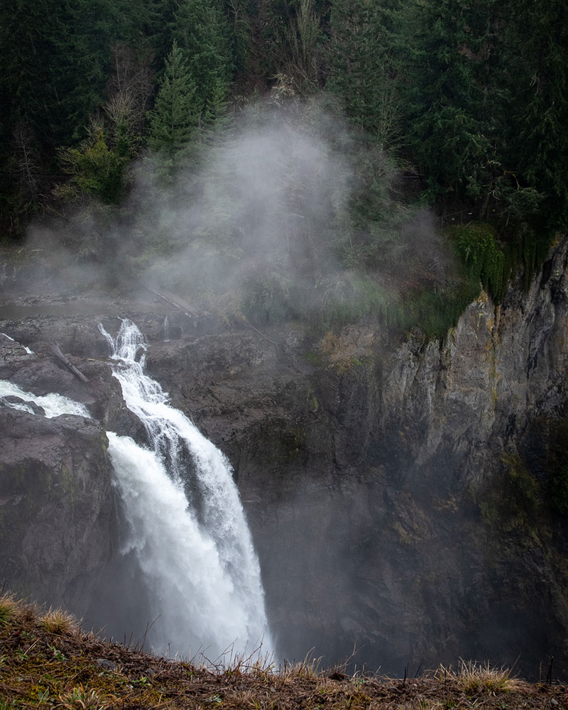 Mist rises from the top of a waterfall, surrounded by cliffs and trees