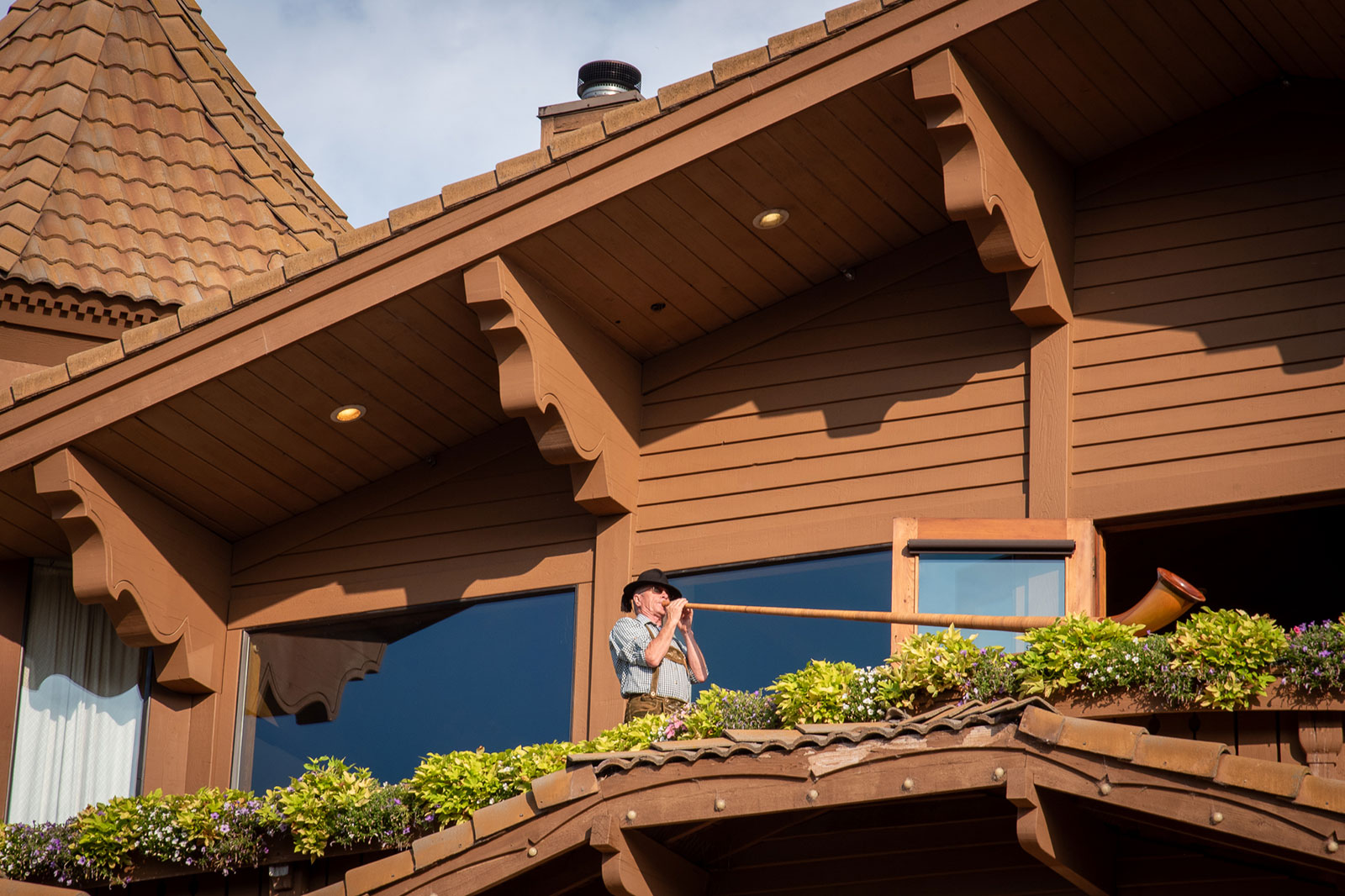 A man in lederhose blows into a large horn on a balcony.