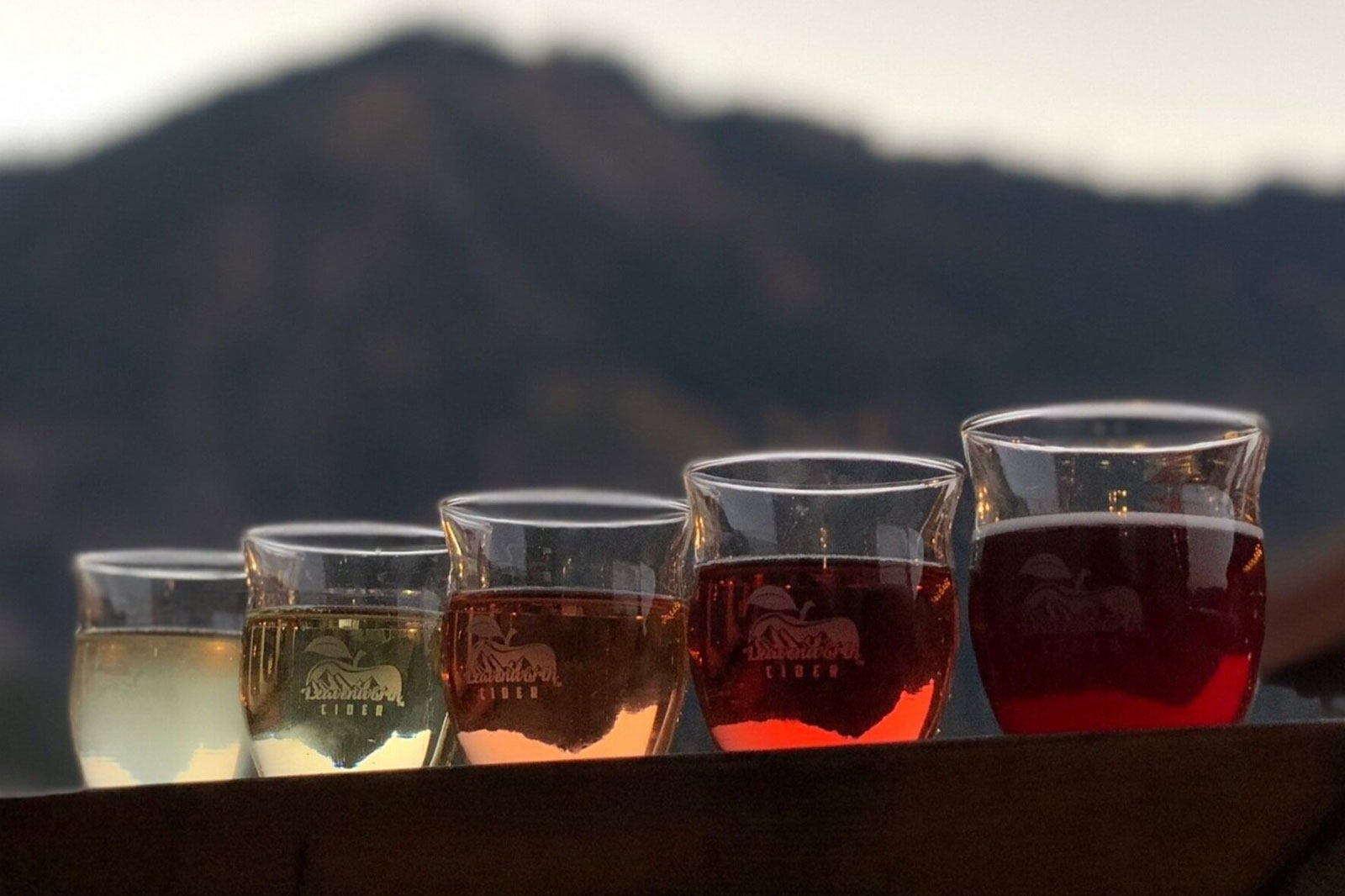 Five glasses have cups of varying colors from light yellow to dark red are seen in front of a mountain range.