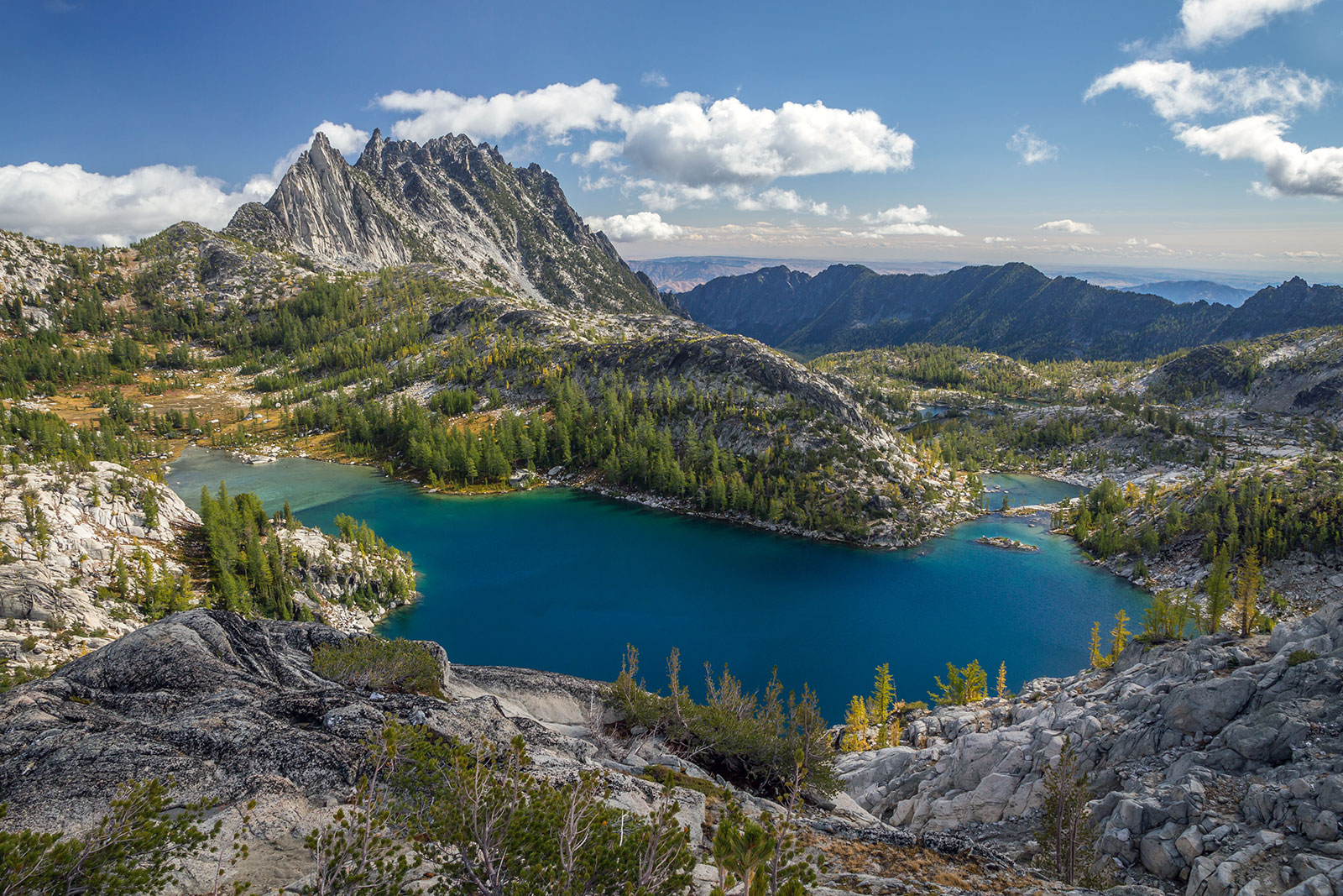 An aerial view of a bright blue lake amind granite rocks, green trees, and blue skies.