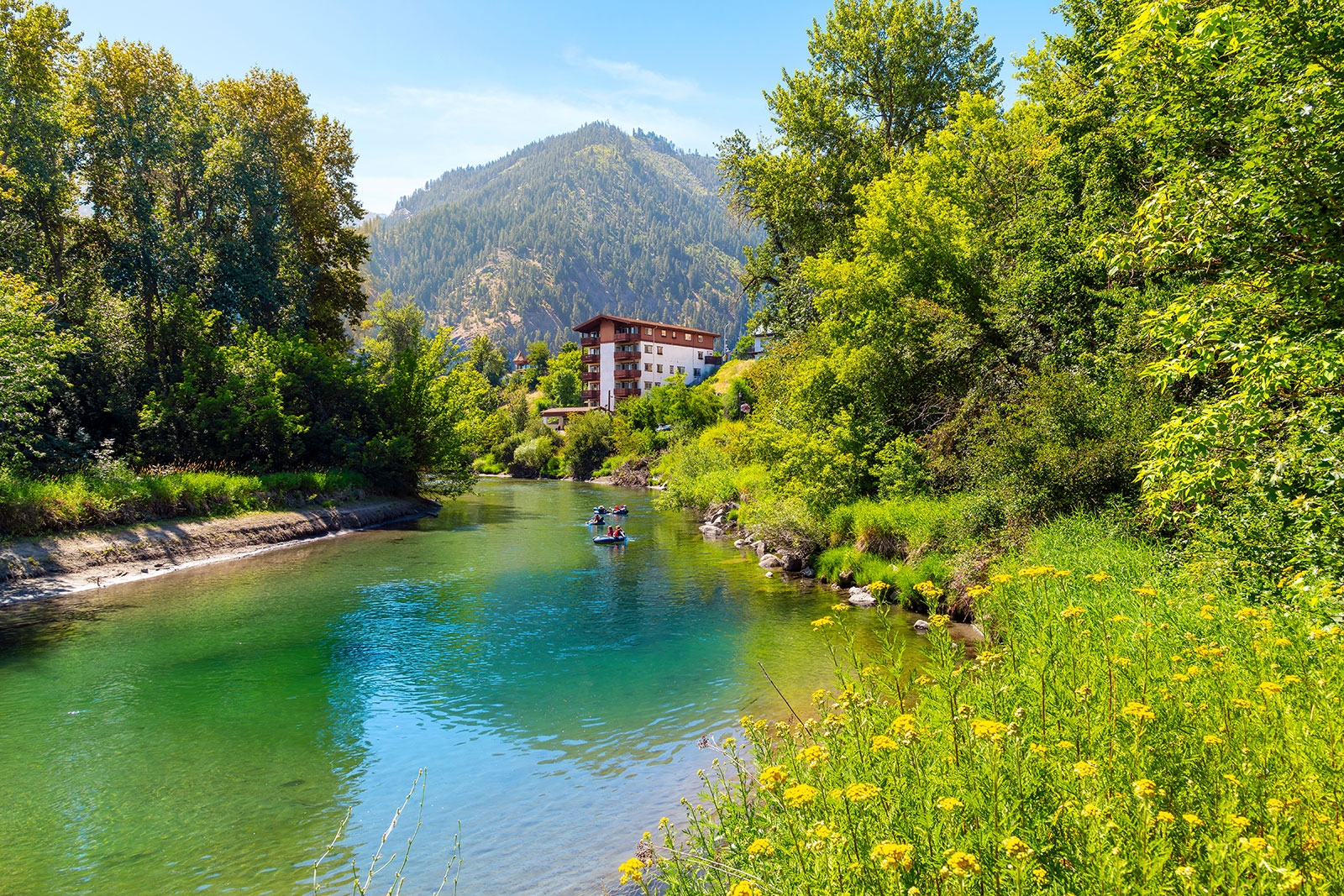 People float down a green-blue river, surrounded by trees and yellow flower. In the background is a hotel and a mountain.