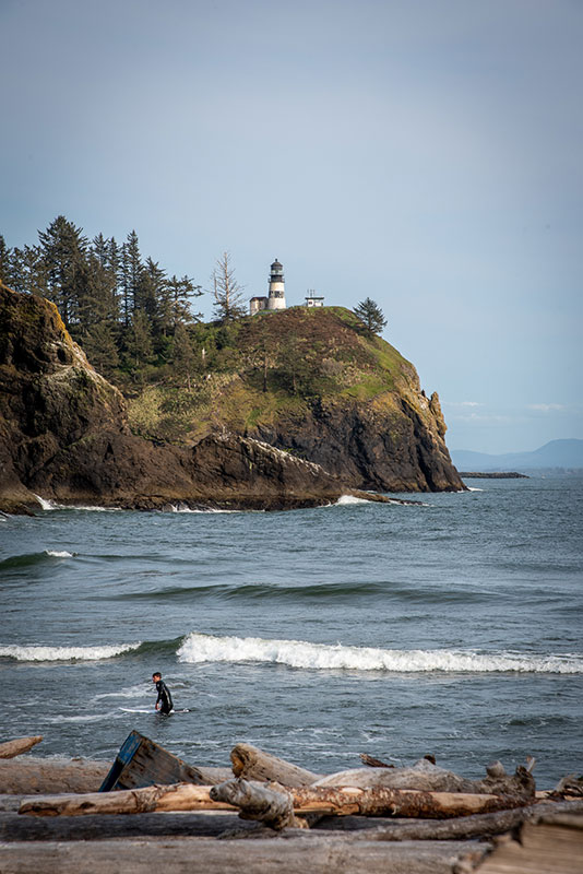 A surfer waits for wave. In the background, Cape Disappointment lighthouse sits above the cliff.