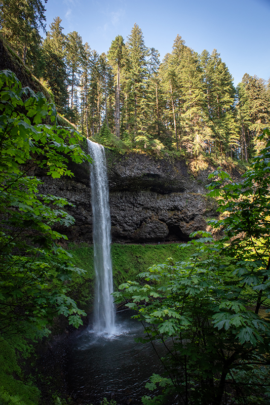 A tall waterfall cascades over rocks to a pool below.